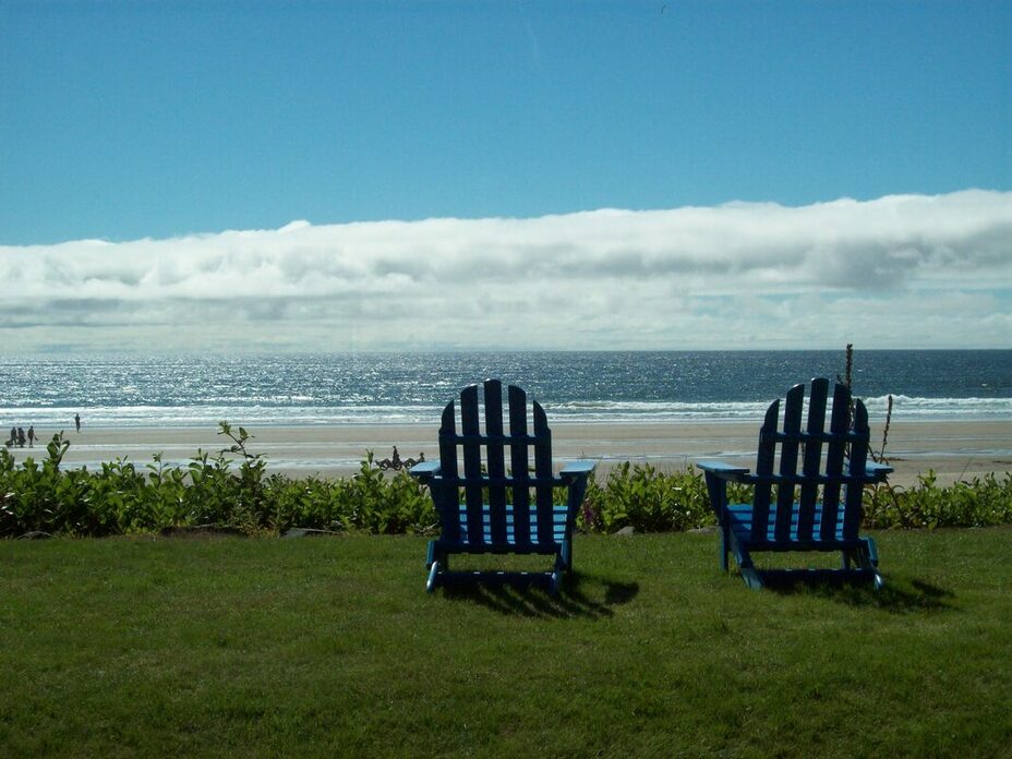 Photograph of two blue wooden deck chairs on a green lawn overlooking a sandy beach, blue ocean with white foamy waves, and a cloudy sky.